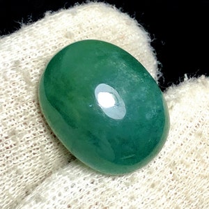 Puede incluir: Una gema pulida, de forma ovalada, en un tono verde bosque profundo. La piedra tiene una superficie lisa y brillante con un brillo sutil, que refleja la luz. La gema se encuentra sobre un fondo blanco texturizado.