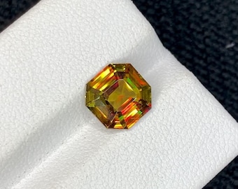May include: A faceted gemstone with an octagonal shape. The gem displays a blend of colors, including yellow, orange, and green, with a central core of deeper hues. The gem is set against a white surface.