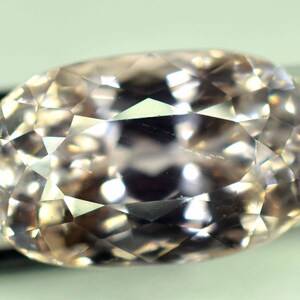 Puede incluir: Una gema facetada, de forma ovalada y color melocot&oacute;n p&aacute;lido. La gema est&aacute; tallada con numerosas facetas peque&ntilde;as, creando un efecto brillante. La piedra est&aacute; sujeta por un par de finas pinzas met&aacute;licas oscuras, sobre un fondo neutro.