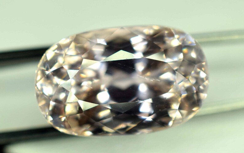 Puede incluir: Una gema facetada, de forma ovalada y color melocot&oacute;n p&aacute;lido. La gema est&aacute; tallada con numerosas facetas peque&ntilde;as, creando un efecto brillante. La piedra est&aacute; sujeta por un par de finas pinzas met&aacute;licas oscuras, sobre un fondo neutro.