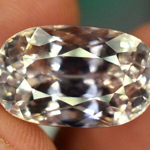 Puede incluir: Una gema facetada de forma ovalada con un color p&aacute;lido y transl&uacute;cido. La gema se sostiene entre los dedos, mostrando su intrincado corte y superficies reflectantes. Las facetas de la gema capturan la luz, creando un efecto brillante.