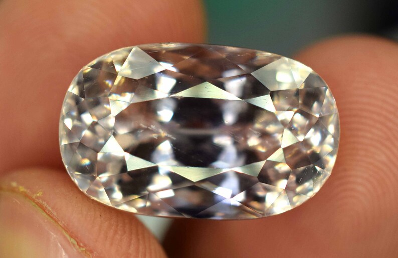 Puede incluir: Una gema facetada de forma ovalada con un color p&aacute;lido y transl&uacute;cido. La gema se sostiene entre los dedos, mostrando su intrincado corte y superficies reflectantes. Las facetas de la gema capturan la luz, creando un efecto brillante.