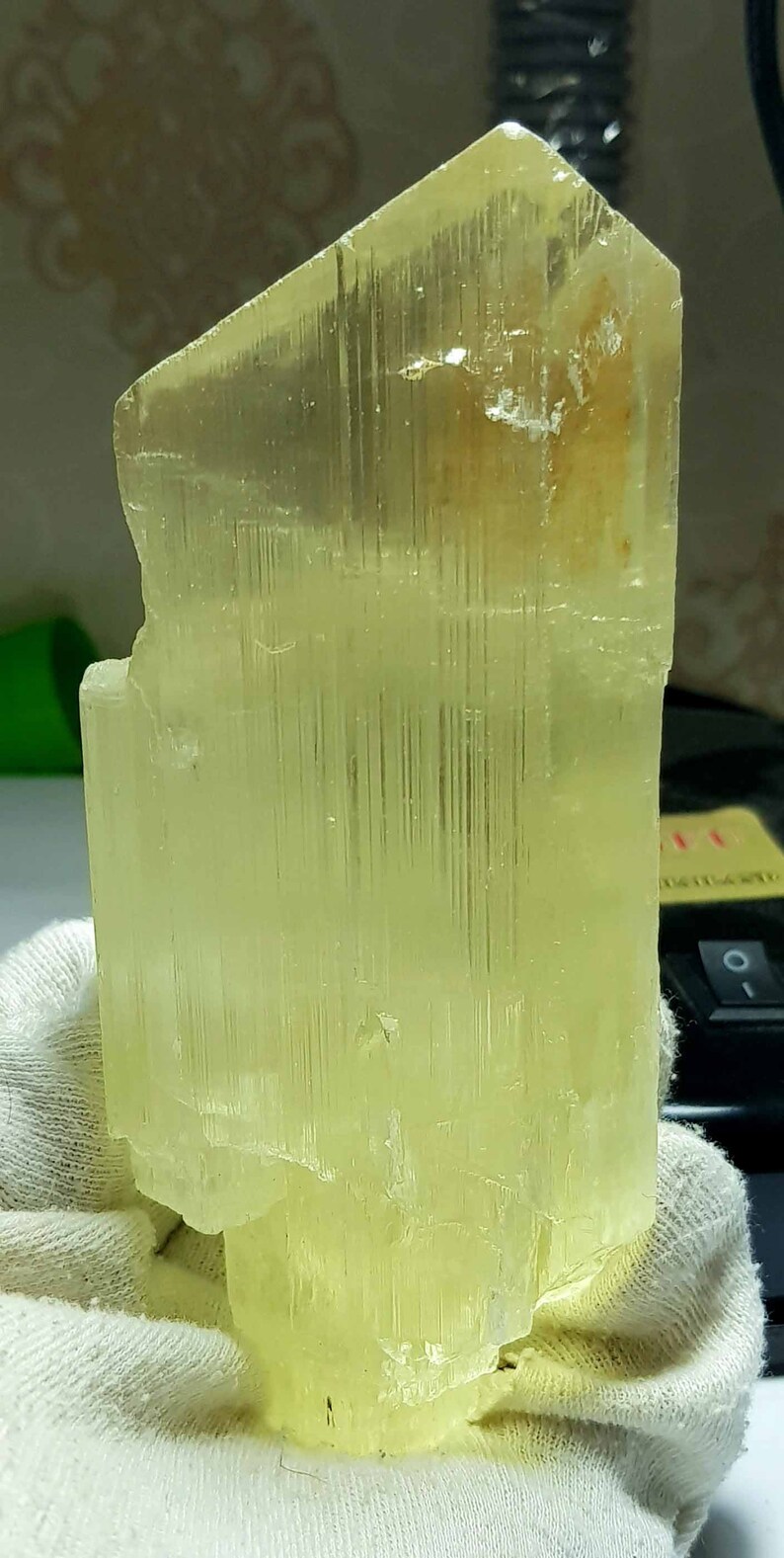 May include: A translucent, pale yellow crystal formation. The mineral has a rectangular shape with vertical striations, and is held by a gloved hand. The crystal's color is consistent throughout, with a slightly rough texture.