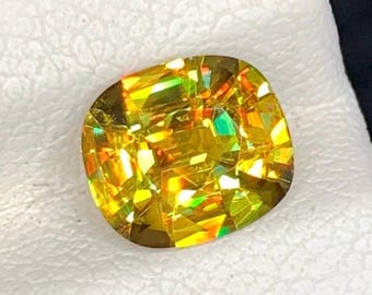May include: A cushion-cut gemstone with vibrant yellow and green hues. The faceted surface reflects light, creating a sparkling effect. The gemstone is set against a white background, highlighting its color and brilliance.