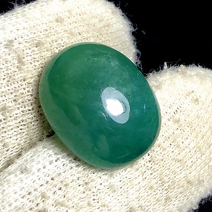 Puede incluir: Una piedra preciosa verde, pulida y de forma ovalada. La piedra tiene una superficie lisa y brillante con un ligero brillo. La piedra preciosa est&aacute; colocada sobre un fondo blanco texturizado, con un fondo oscuro.