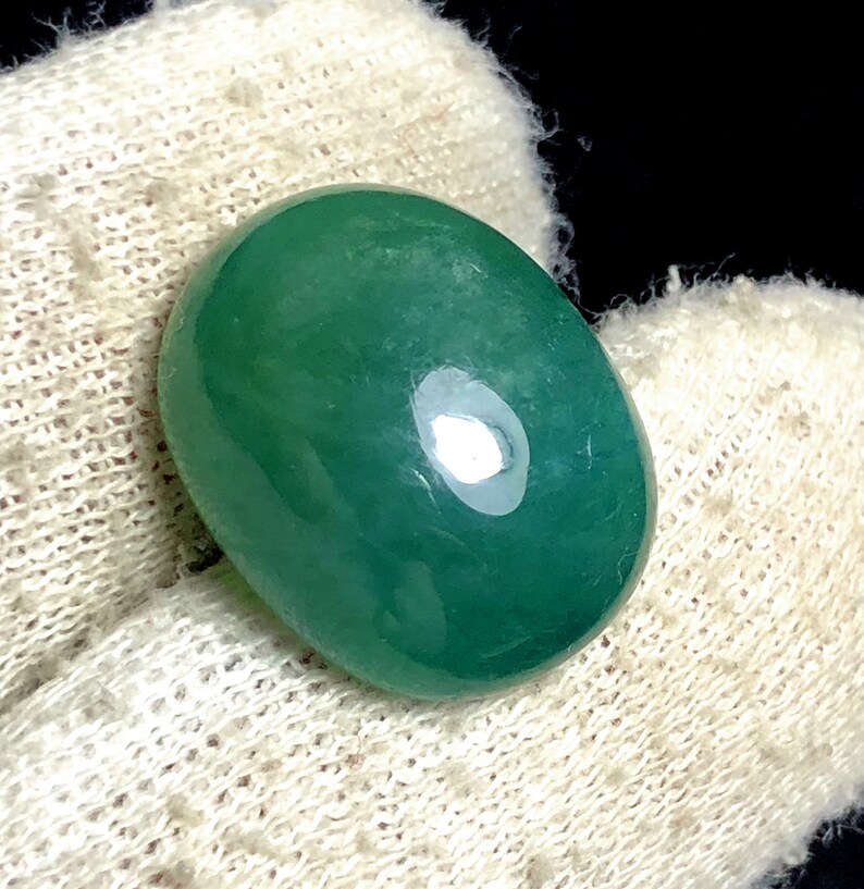 Puede incluir: Una piedra preciosa verde, pulida y de forma ovalada. La piedra tiene una superficie lisa y brillante con un ligero brillo. La piedra preciosa est&aacute; colocada sobre un fondo blanco texturizado, con un fondo oscuro.