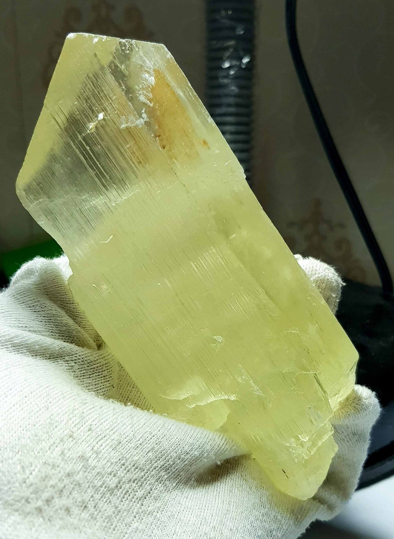 May include: A large, pale yellow crystal held in a white gloved hand. The crystal has a rectangular shape with a faceted surface and a slightly rough texture. The color is a soft, translucent yellow.