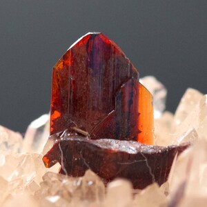 May include: Close-up of a mineral specimen featuring a cluster of reddish-brown crystals. The crystals have a glossy, translucent appearance with hints of orange and purple. The base is composed of smaller, lighter-colored crystals.