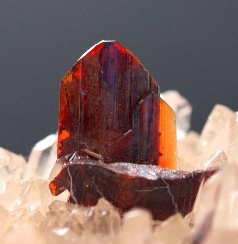 May include: Close-up of a mineral specimen featuring a cluster of reddish-brown crystals. The crystals have a glossy, translucent appearance with hints of orange and purple. The base is composed of smaller, lighter-colored crystals.