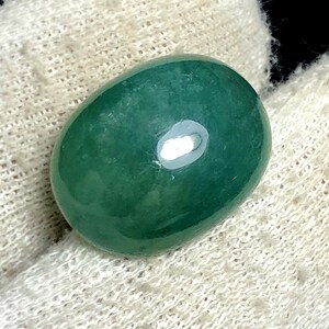 Puede incluir: Una gema ovalada pulida, de un tono verde intenso y rico. La piedra tiene una superficie lisa y brillante con un brillo sutil, que refleja la luz. La gema se encuentra sobre un fondo blanco texturizado.