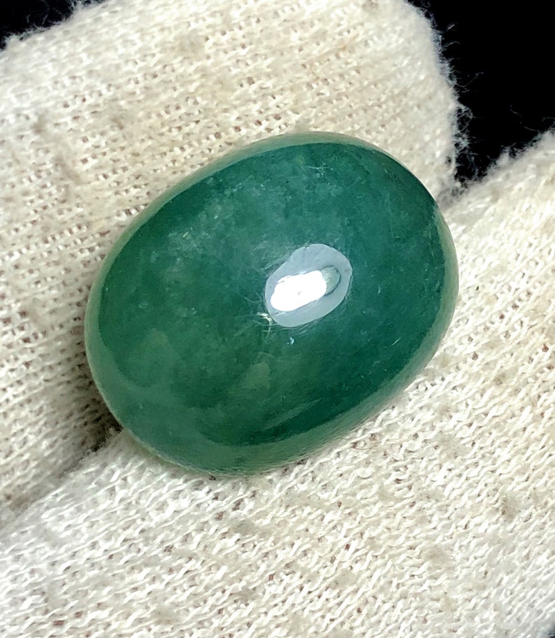Puede incluir: Una gema ovalada pulida, de un tono verde intenso y rico. La piedra tiene una superficie lisa y brillante con un brillo sutil, que refleja la luz. La gema se encuentra sobre un fondo blanco texturizado.