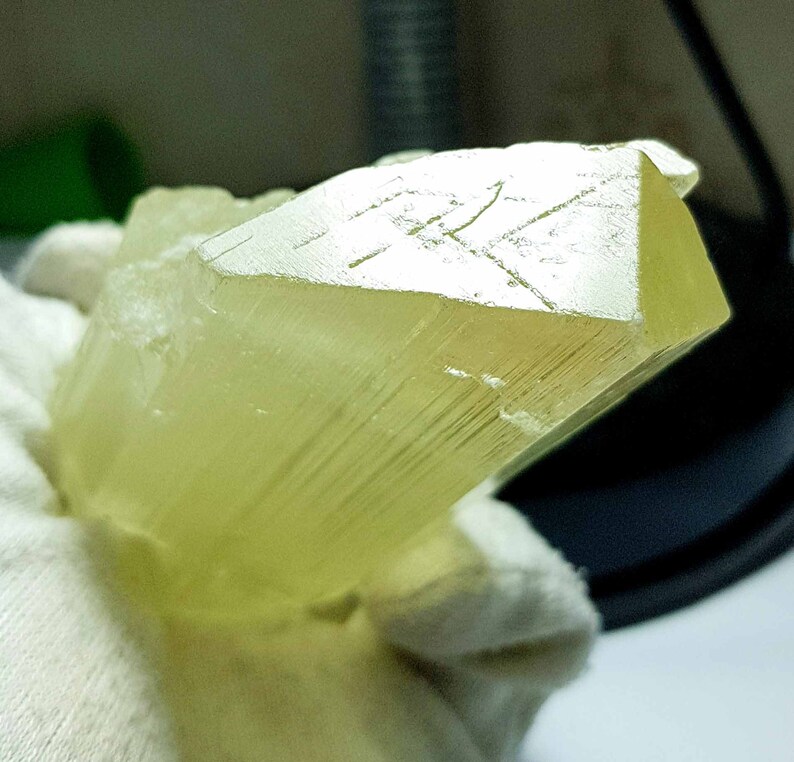 May include: A translucent, pale yellow crystal formation with a geometric, faceted end. The crystal has a rectangular shape with visible striations along its length. The overall appearance is smooth and polished, with a subtle glow.