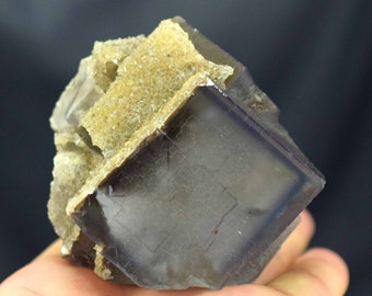 May include: A close-up of a fluorite mineral specimen. The cube-shaped crystal has a dark purple hue with a lighter purple border. The top of the specimen has a light tan, crystalline structure.