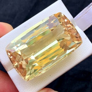Könnte beinhalten: Ein großer, rechteckiger Edelstein in warmen Gelb- und Goldtönen. Der Stein ist facettiert, wodurch Licht reflektiert wird. Er liegt auf einer weißen Oberfläche mit einem weißen Band vor dunklem Hintergrund.