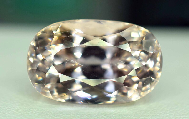 Puede incluir: Una gema facetada de forma ovalada con un tono melocot&oacute;n p&aacute;lido. La superficie de la gema est&aacute; dividida en numerosas facetas triangulares peque&ntilde;as, creando un efecto brillante. La piedra se encuentra sobre un fondo suave y neutro.