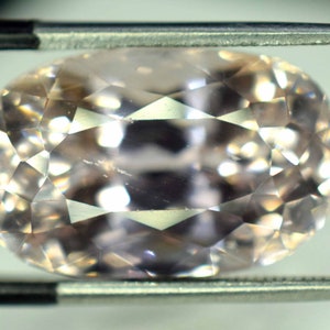 Puede incluir: Una gema facetada de forma ovalada sujeta con pinzas. La gema es de color melocot&oacute;n p&aacute;lido y tiene un tallado brillante, que refleja la luz en sus m&uacute;ltiples facetas. Las pinzas son de color plateado.