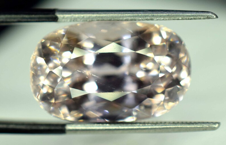 Puede incluir: Una gema facetada de forma ovalada sujeta con pinzas. La gema es de color melocot&oacute;n p&aacute;lido y tiene un tallado brillante, que refleja la luz en sus m&uacute;ltiples facetas. Las pinzas son de color plateado.