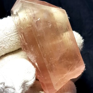 K&ouml;nnte beinhalten: Ein durchscheinender, pfirsichfarbener Kristall mit rechteckiger Form. Der Kristall hat eine glatte, polierte Oberfl&auml;che mit sichtbaren inneren Strukturen. Der Kristall wird in einem wei&szlig;en Handschuh vor dunklem Hintergrund gehalten.