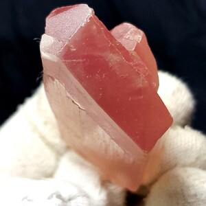 K&ouml;nnte beinhalten: Ein durchscheinender, rot-rosa Kristallcluster. Der Kristall hat eine facettierte, geometrische Form mit einer leicht rauen Textur. Die Farbe ist durchgehend, mit einem weichen, leuchtenden Aussehen. Der Kristall wird vor einem wei&szlig;en Hintergrund gehalten.