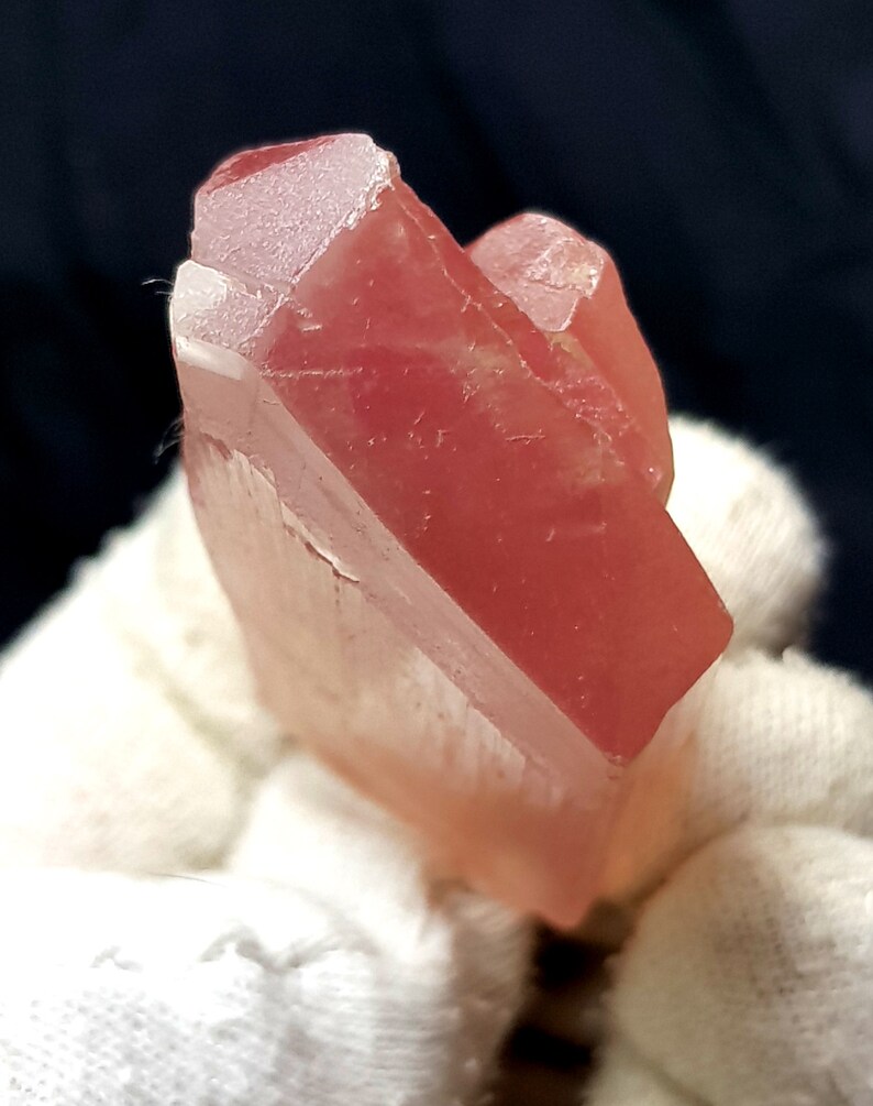 K&ouml;nnte beinhalten: Ein durchscheinender, rot-rosa Kristallcluster. Der Kristall hat eine facettierte, geometrische Form mit einer leicht rauen Textur. Die Farbe ist durchgehend, mit einem weichen, leuchtenden Aussehen. Der Kristall wird vor einem wei&szlig;en Hintergrund gehalten.