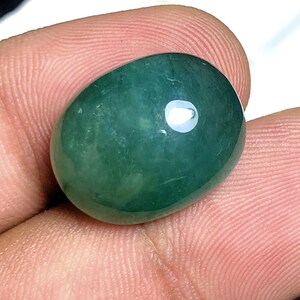 Puede incluir: Una gema pulida, de forma ovalada, de color verde azulado intenso. La piedra tiene una superficie lisa y brillante con un punto brillante y reflectante. La gema se sostiene entre los dedos, mostrando su tama&ntilde;o y forma. Adecuada para la fabricaci&oacute;n de joyas.
