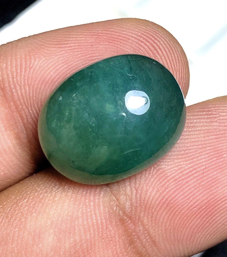 Puede incluir: Una gema pulida, de forma ovalada, de color verde azulado intenso. La piedra tiene una superficie lisa y brillante con un punto brillante y reflectante. La gema se sostiene entre los dedos, mostrando su tama&ntilde;o y forma. Adecuada para la fabricaci&oacute;n de joyas.