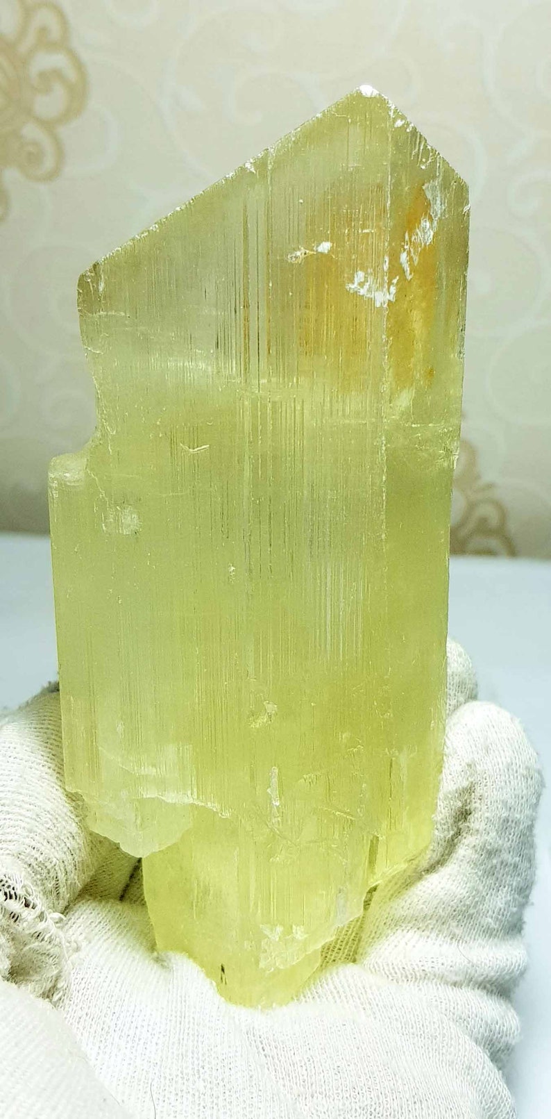 May include: A translucent, pale yellow crystal formation. The crystal has a rectangular shape with a slightly angled top and visible vertical striations. The crystal is held by a gloved hand.