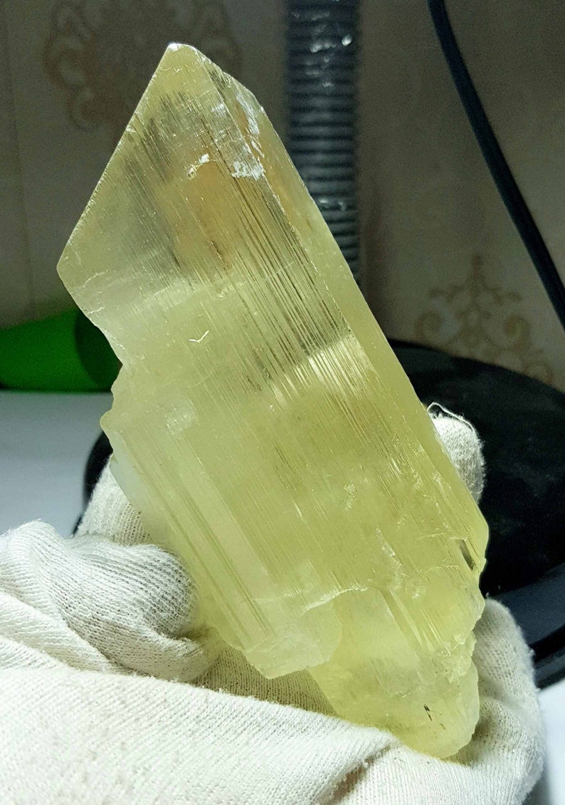 May include: A translucent, pale yellow crystal with a rectangular shape and a textured surface. The crystal is held in a white glove. The crystal's facets catch the light, revealing internal striations. This mineral specimen is a beautiful example of natural crystal formations.