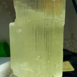 May include: A translucent, pale yellow crystal with a rectangular shape. The crystal has vertical striations and a slightly rough texture. The object is held by a gloved hand, suggesting it is a mineral specimen.