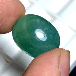 Puede incluir: Una gema verde pulida de forma ovalada. La gema se sostiene entre dos dedos, mostrando su superficie lisa y reflectante. La piedra tiene una ligera translucidez, con una sutil variaci&oacute;n de color.