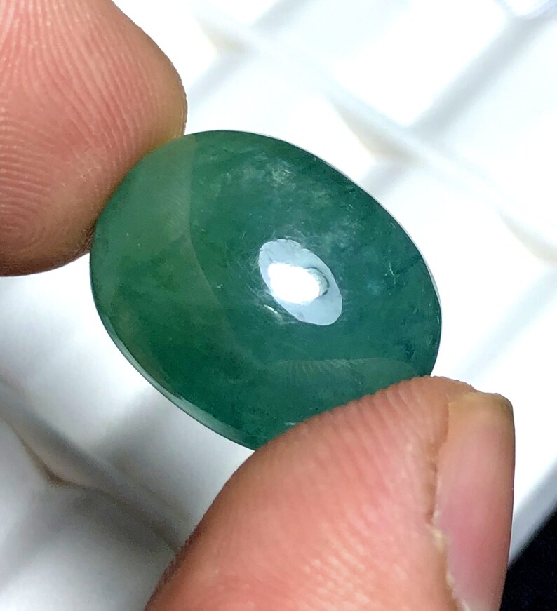 Puede incluir: Una gema verde pulida de forma ovalada. La gema se sostiene entre dos dedos, mostrando su superficie lisa y reflectante. La piedra tiene una ligera translucidez, con una sutil variaci&oacute;n de color.