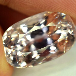 Puede incluir: Primer plano de una gema facetada de forma ovalada. La gema es de color melocot&oacute;n p&aacute;lido con inclusiones internas y un alto grado de claridad. Las facetas reflejan la luz, creando un efecto brillante. La gema se sostiene entre dos dedos.