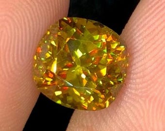 May include: A faceted, cushion-cut gemstone with a vibrant yellow-green hue. The gem displays internal fire with flashes of orange and yellow. The stone is held between two fingers against a dark background, highlighting its brilliance.