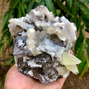 以下が含まれることがあります： グレー、白、淡い黄色が混ざった天然鉱物標本。結晶はざらざらとした質感で、層状に配置されています。標本は手で持たれています。