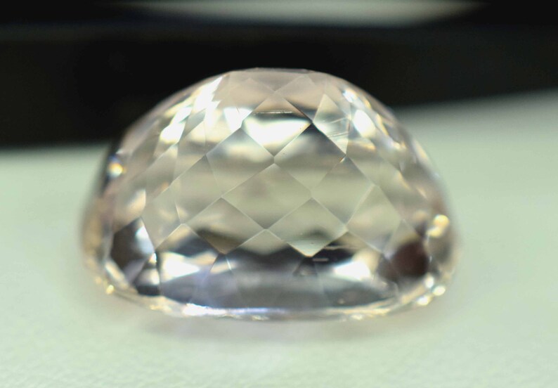 Puede incluir: Una gema facetada de color melocot&oacute;n claro, con una parte superior redondeada. La superficie est&aacute; tallada con numerosas facetas peque&ntilde;as en forma de diamante, creando un efecto brillante. La gema se encuentra sobre un fondo suave y neutro.