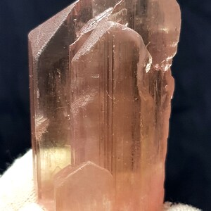 K&ouml;nnte beinhalten: Eine durchscheinende, pfirsichfarbene Kristallformation mit prismatischer Struktur. Der Kristall hat eine raue, strukturierte Oberfl&auml;che und ein nat&uuml;rliches, unpoliertes Aussehen. Die Farbe variiert leicht, wobei einige Bereiche blasser erscheinen.