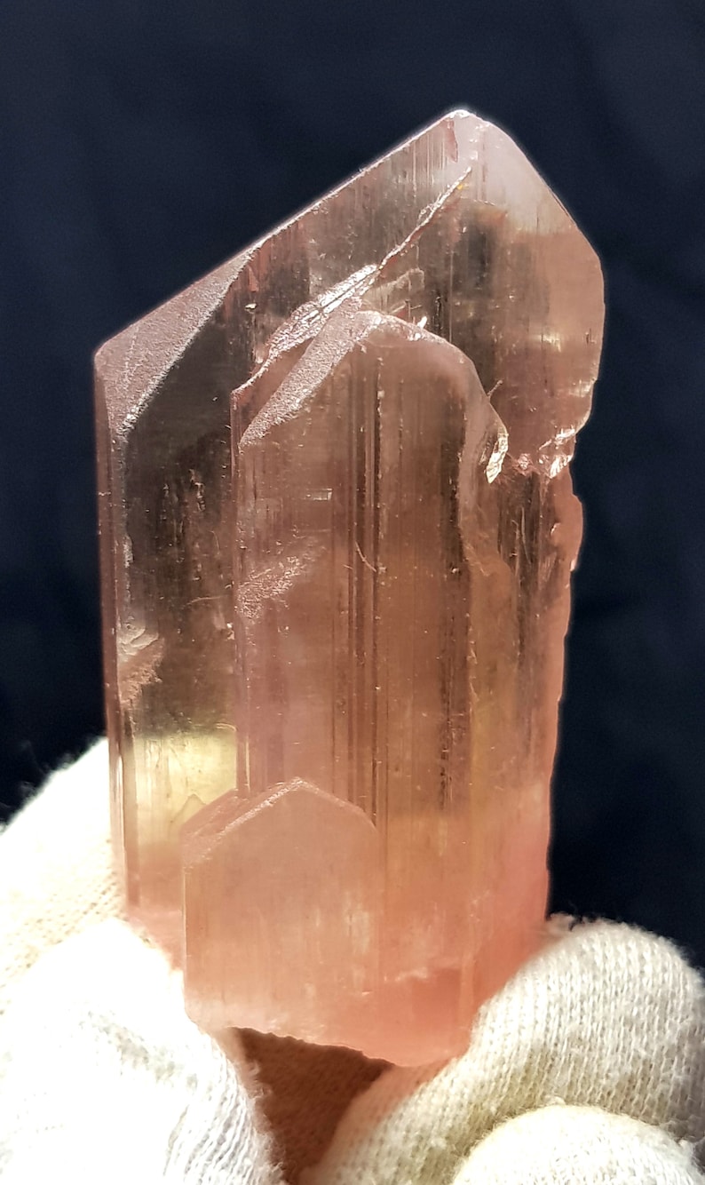K&ouml;nnte beinhalten: Eine durchscheinende, pfirsichfarbene Kristallformation mit prismatischer Struktur. Der Kristall hat eine raue, strukturierte Oberfl&auml;che und ein nat&uuml;rliches, unpoliertes Aussehen. Die Farbe variiert leicht, wobei einige Bereiche blasser erscheinen.