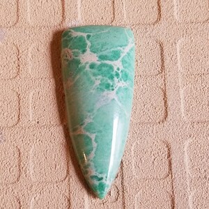May include: A green and white cabochon gemstone with a pointed shape. The stone has a marbled pattern with white veins and green swirls.
