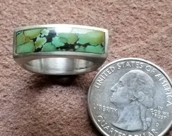 Puede incluir: Un anillo de plata con una incrustación de turquesa verde. El anillo está al lado de una moneda de veinticinco centavos de dólar estadounidense para comparar el tamaño.