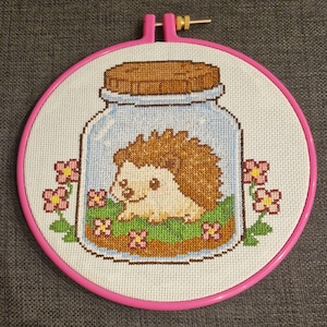 May include: A cross-stitch embroidery featuring a hedgehog inside a jar, surrounded by flowers. The hedgehog has brown quills and a cream-colored face. The embroidery is set in a pink hoop.