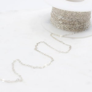 Peut inclure: Une bobine de chaîne argentée, avec une partie de la chaîne déroulée et posée sur une surface blanche.