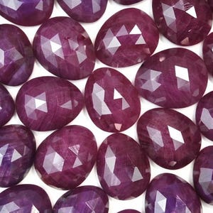 May include: A collection of faceted ruby gemstones, each with a unique, irregular shape. The rubies are a deep, rich red color and have a polished, shiny finish.