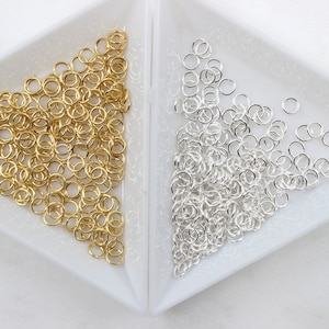 May include: A close-up of two piles of jump rings, one gold and one silver. The jump rings are small and round, and they are arranged in a random pattern.