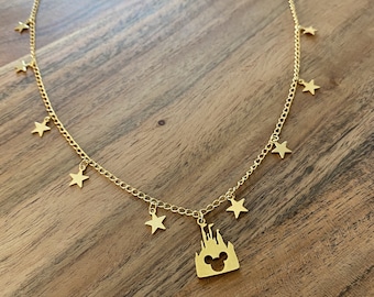 May include: A gold necklace with a charm of a castle with Mickey Mouse ears cut out. The necklace has a chain with small star charms hanging from it.