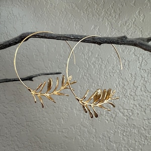 May include: A pair of gold-colored hoop earrings. Each earring features a delicate branch design with detailed leaves. The earrings are suspended from a dark, textured branch against a neutral background.