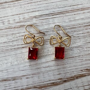 May include: Gold bow earrings with red square-shaped gemstones dangling from the bows.