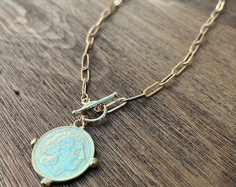Puede incluir: Un collar de cadena dorada con un gran colgante de moneda de oro. La moneda tiene el perfil de una persona.