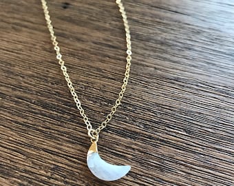 May include: A delicate gold-colored chain necklace featuring a crescent moon pendant. The moon pendant is a light blue color with a gold-colored cap. The necklace is displayed on a wooden surface.