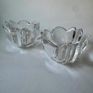 May include: Two clear glass bowls with a flower petal design. The bowls are made of transparent glass and have a wavy, scalloped edge. The bowls are empty and sit on a white surface.