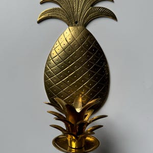 May include: A gold pineapple-shaped wall sconce with a candle holder. The sconce has a detailed pineapple design with a textured surface.